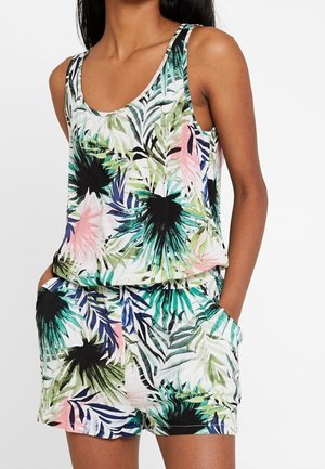 Vrouw draagt een mouwloze tropicalprint romper met groene, zwarte, roze en blauwe bladerenpatronen en haar handen in de zakken.