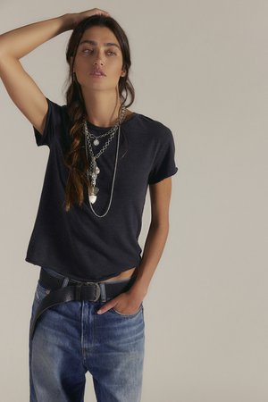 Kobieta z długim warkoczem, ubrana w czarny t-shirt, warstwowe srebrne naszyjniki, niebieskie dżinsy i czarny pasek, pozująca z jedną ręką na głowie.
