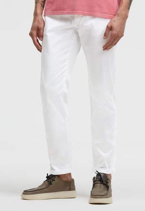 Mann trägt hochgekrempelte weiße Hosen, braune Schnürschuhe und ein rosa Hemd vor einfachem hellem Hintergrund.