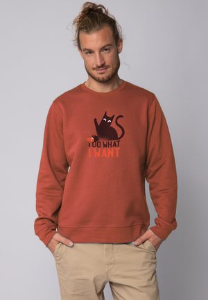 Roestkleurige sweatshirt gemaakt van zachte stof. Bevat een afbeelding van een zwarte kat met de tekst "IK DOE WAT IK WIL" in contrasterend oranje. Eenvoudig ontwerp met ronde hals.
