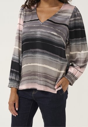 Vrouw draagt een blouse met lange mouwen en V-hals met horizontale aquarelstrepen in grijstinten, zwart en roze, gecombineerd met donkerblauwe jeans.