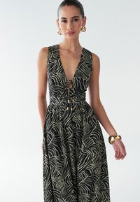 Vestido negro con escote en V profundo y cintura ceñida, con un patrón abstracto ondulado en crema. Pendientes y brazalete dorados llamativos completan el conjunto.