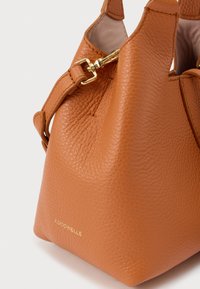 Καφέ τσάντα χειρός από δέρμα με υφή, με χρυσό κούμπωμα και το λογότυπο "Coccinelle" στο μπροστινό μέρος, εμφανίζοντας μέρος του λουριού και το ανοιχτό πάνω μέρος.