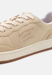 Beige suede sneaker with textured surface, white laces, and a cream midsole. Features embossed text "ANYTHING CAN BE CHANGED" on the side.