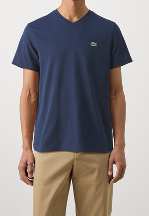 Mężczyzna ubrany w granatowy T-shirt z dekoltem w serek i małym zielonym logo krokodyla na lewej piersi oraz beżowe spodnie.