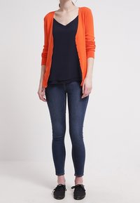Cardigan arancione sopra una maglietta a V blu navy, abbinato a jeans skinny blu scuro e sneakers nere. Design semplice e silhouette aderente.