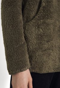 Olivengrøn, fuzzy fleece-overdel med en struktureret overflade. Har en rund skjortekant og lange ærmer, parret med ribbede sorte bukser.