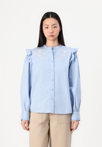 Chemise bleu clair à boutons avec des épaules froncées, broderies florales sur la poitrine et poignets froncés, fabriquée à partir d'un tissu texturé.