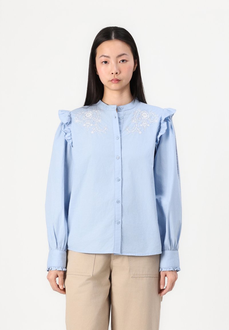 Chemise bleu clair à boutons avec des épaules froncées, broderies florales sur la poitrine et poignets froncés, fabriquée à partir d'un tissu texturé.