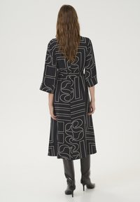 Abito midi nero con motivi astratti bianchi, maniche lunghe e cintura da annodare in vita. Presenta un design fluido e una texture liscia. Abbinato a stivali alti fino al ginocchio.