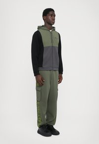 Sweatshirt com capuz com fecho éclair em verde e cinza. Calças cargo em oliva com detalhes em padrão verde. Ténis pretos. Textura de tecido suave.