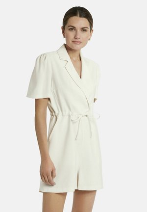 Vrouw met donker haar in een staart, gekleed in een witte korte mouwen romper met kraag en tailleband, staand tegen een effen lichte achtergrond.