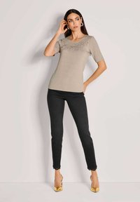 Beige Oberteil mit kurzen Ärmeln und dekorativen metallischen Akzenten auf der Brust, kombiniert mit schwarzen Skinny-Jeans und goldenen High-Heels.