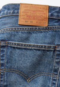 Jean en denim bleu avec une patch en cuir tan comportant un texte en relief, des détails de couture, et un design classique de poche arrière. Taille W32 L32.