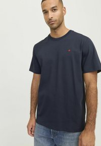 Camiseta de algodón azul marino con mangas cortas, que presenta un pequeño logo de punto rojo en el pecho. Corte casual, cuello redondo y textura suave.