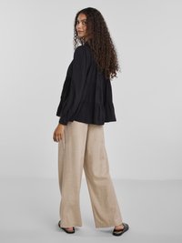Kvinne med langt krøllete hår iført en løs svart bluse, beige vide bukser og svarte flate sko, stående mot en enkel lys bakgrunn.