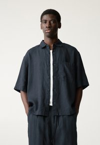 Camicia corta blu scuro in lino, con frontale a bottoni, tasca sul petto e vestibilità morbida. Indossata sopra un top bianco.