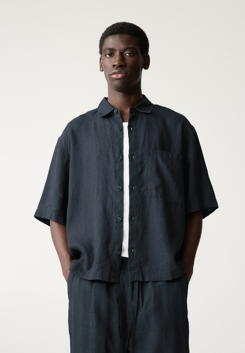 Chemise à manches courtes bleu marine foncé en lin, avec un devant à boutons, une poche sur la poitrine et une coupe décontractée. Portée par-dessus un débardeur blanc.