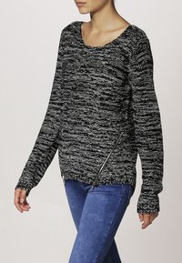 Schwarzer und weißer strukturierter Pullover mit weitem Ausschnitt, langen Ärmeln und seitlichem Reißverschluss, kombiniert mit blauen Jeans.