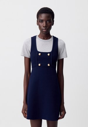 Abito navy con corsetto a coste, scollo quadrato e accenti di bottoni dorati, indossato sopra una maglia grigia a maniche corte. Tessuto liscio; design aderente.