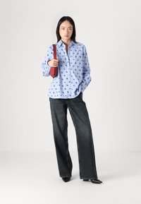 Light blue button-up shirt with black patterns, paired with wide-leg dark jeans, and a red shoulder bag. Black pointed shoes complete the look.