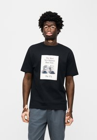 Μαύρο βαμβακερό t-shirt με γραφικό τύπωμα από αθλητικά παπούτσια και κείμενο από πάνω. Το μοντέλο φορά επίσης μπλε-γκρι παντελόνια, στέκεται μπροστά σε λευκό φόντο.