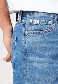 Světle modré džínové kalhoty s klasickým střihem, s bílou cK nášivkou na zadní kapse a stříbrnými kovovými detaily. Viditelné švy.