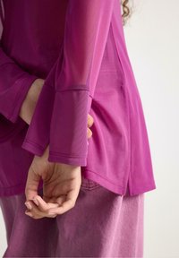 Langærmet top i klart pink stof med en glat tekstur. Har vide ærmer og en afslappet pasform med sideslids nær kanten.