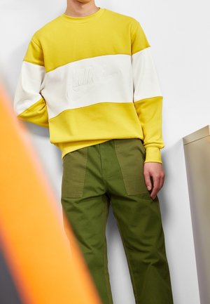 Persoon in een geel en wit Nike-sweater, gecombineerd met olijfgroene broek, staande tegen een witte muur met een vervaagd oranje object in de buurt.