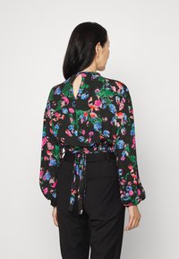 Blusa preta com padrão floral vibrante em rosa, azul e verde, mangas longas e costas com recorte em forma de lágrima e detalhe de laço. Combine com calças escuras.