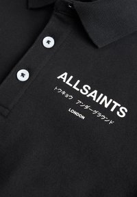 Fekete poloing, amely texturált anyagból készült. A galléron három fehér gomb található, az "ALLSAINTS" logóval és további fehér felirattal.