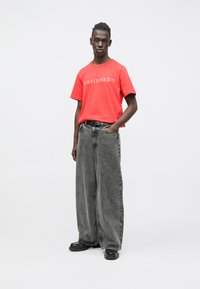 T-shirt vermelha de algodão com estampa "marimekko", combinada com jeans cinzentos largos e folgados. A modelo usa sapatos pretos e um cinto preto.