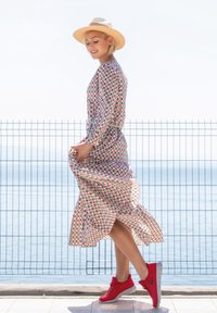 Robe à manches longues à motifs avec des designs géométriques rouges et verts, nouée à la taille. Associée à des chaussures de sport rouges et un chapeau en paille.