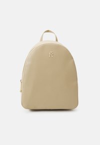 Zaino beige con parte superiore curva, texture liscia simile alla pelle, dettaglio logo dorato e manico superiore. Chiusura a zip su un lato.