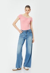 Top corto a maniche corte a righe rosa con dettaglio arricciato, abbinato a jeans a gamba larga blu chiaro a vita alta. Il modello indossa sneaker bianche e marroni.