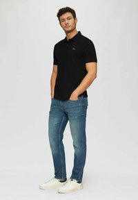 Schwarzes Poloshirt mit Kragen, kurzen Ärmeln und einem goldenen Logo auf der linken Brust; kombiniert mit engen blauen Jeans und weißen Sneakern.