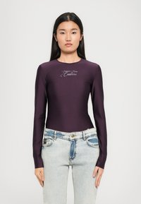Dlhý rukáv body suit v hlbokofialovej látke s hladkou textúrou, s vyšívaným logom "Couture" v svetlej farbe, v kombinácii so svetlomodrými džínami.