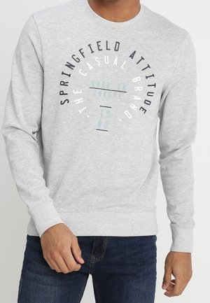 Hombre con sudadera gris claro con texto circular que dice "SPRINGFIELD ATTITUDE THE CASUAL BRAND" y vaqueros azul oscuro.