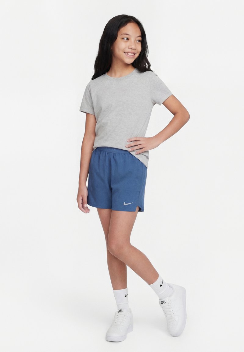 Κορίτσι που στέκεται με το χέρι στη μέση, φοράει γκρι μπλουζάκι, μπλε σορτς Nike, λευκές κάλτσες Nike και λευκά αθλητικά παπούτσια, χαμογελά και κοιτάζει δεξιά.