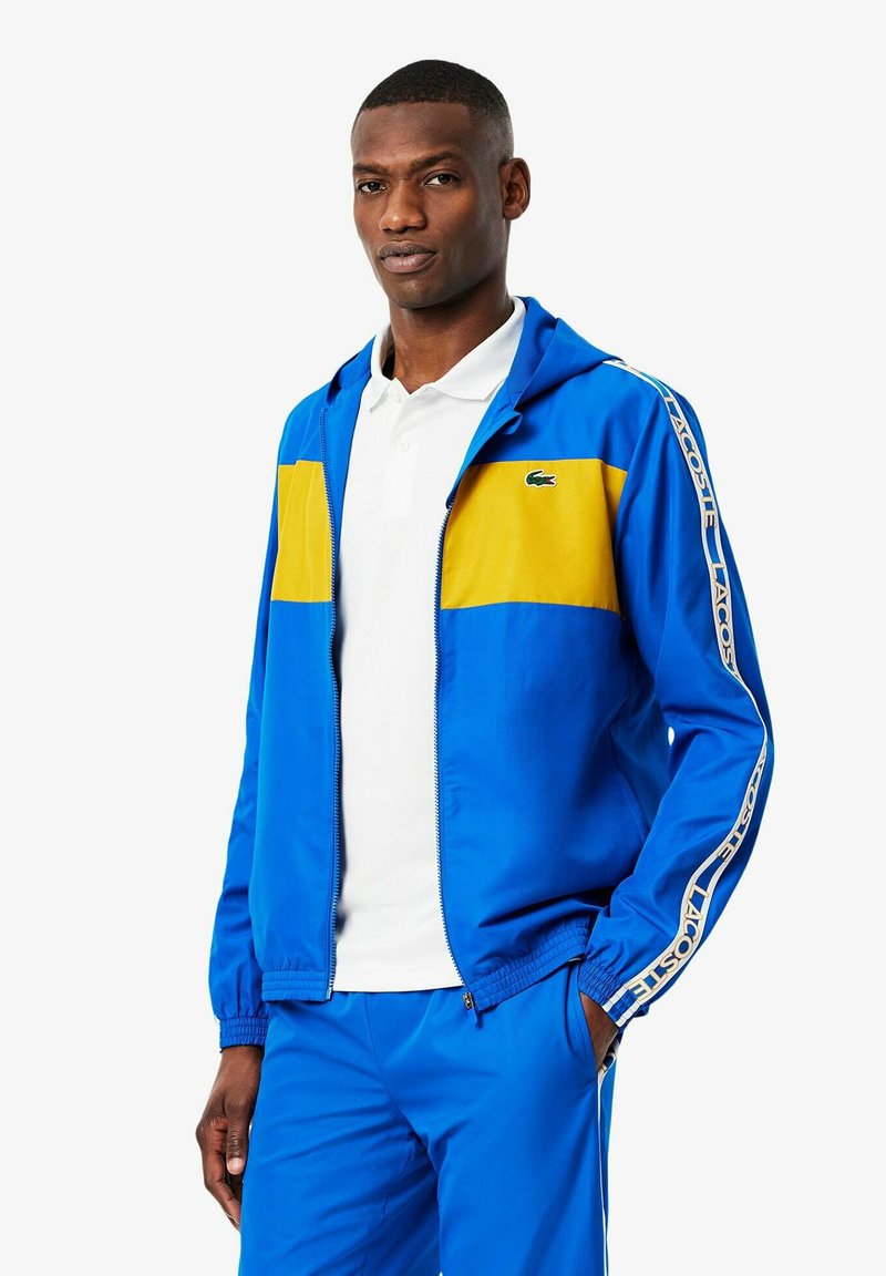 Blauwe trainingsjack met gele panelen; heeft een capuchon, ritssluiting en een kenmerkend logo. Gladde textuur, gecombineerd met een witte polo.