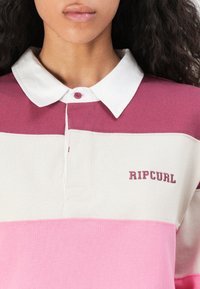 Kvinne iført en rugbyshirt med lange ermer, hvit krage og horisontale striper i maroon, krem og rosa, "RIP CURL"-logo på brystet.