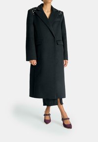 Lungo cappotto nero in lana, con dettagli borchiati sulle spalle, collo a bavero, due tasche frontali e una vestibilità sartoriale.