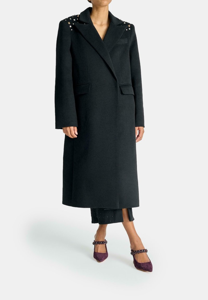 Lungo cappotto nero in lana, con dettagli borchiati sulle spalle, collo a bavero, due tasche frontali e una vestibilità sartoriale.