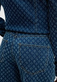 Tela de mezclilla con un pequeño patrón floral en azul claro, mostrando el bolsillo trasero y parte de una camisa de manga larga metida en unos pantalones a juego.
