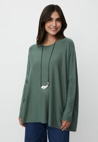 Übergroßer grüner Strickpullover mit langen Ärmeln, der ein seitliches Schlitzdesign aufweist, kombiniert mit einer schwarzen Schnur und einer silbernen Anhänger-Halskette.