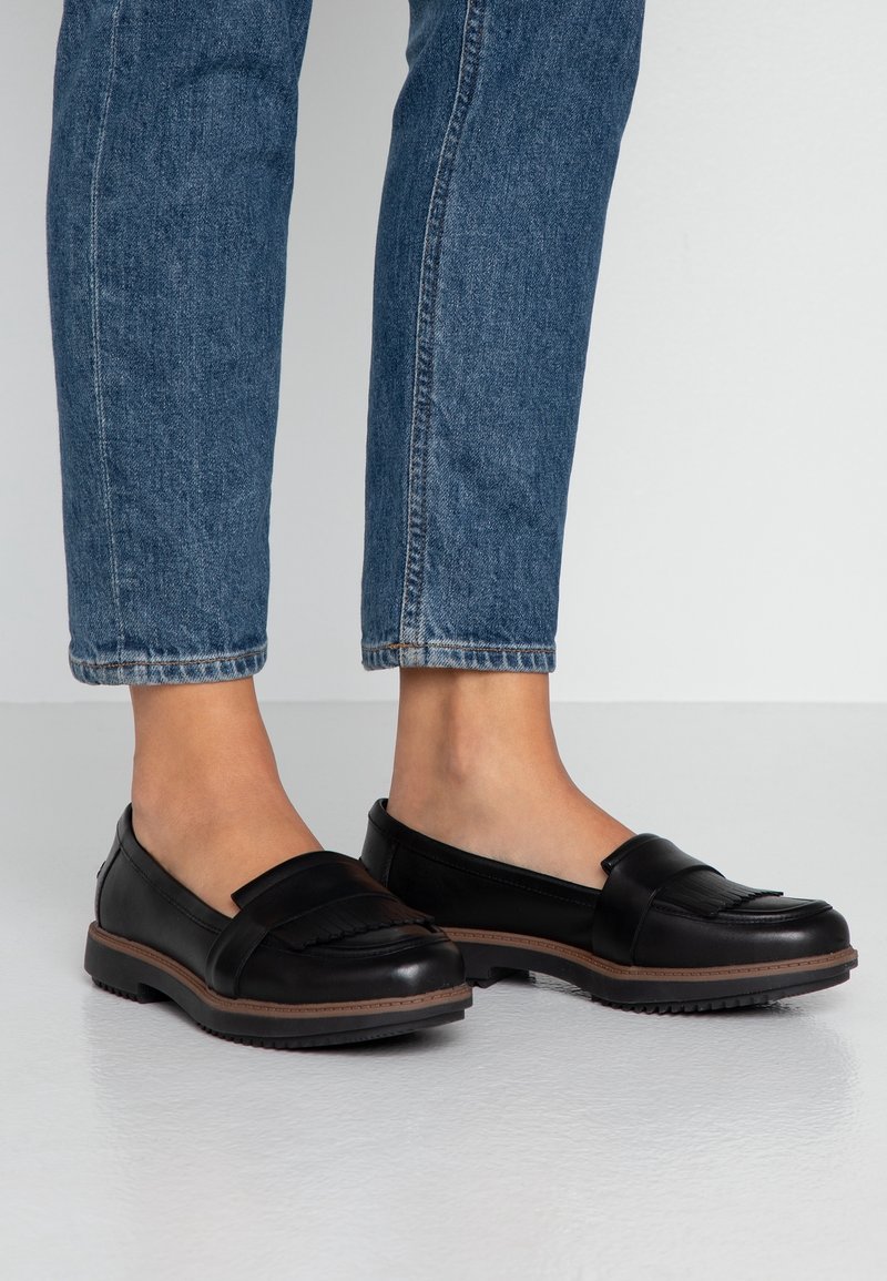 Mocassins en cuir noir avec une tige texturée et un accent frangé, dotés d'une semelle en caoutchouc robuste et de détails marron. Portés avec un jean bleu.