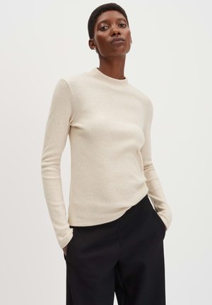Beige, langärmliger Pullover mit hohem Kragen, weicher Textur, figurbetonendem Schnitt, kombiniert mit schwarzen, lässigen Hosen, Hände in den Taschen.