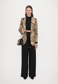 Juostuota zebra raštu blazer iš auksinės ir juodos medžiagos, prigludusi forma, su juodomis plačiomis kelnėmis ir mažai juodai ruda rankine.