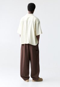 Przesadnie duża żółta koszula w paski z krótkim rękawem i kołnierzykiem, połączona z luźnymi brązowymi spodniami; miękka tekstura i luźny krój; minimalistyczny styl.