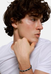 Bracciale in argento con anelli intrecciati, indossato a un polso. L'individuo ha capelli scuri e ricci ed è vestito con una maglietta bianca.